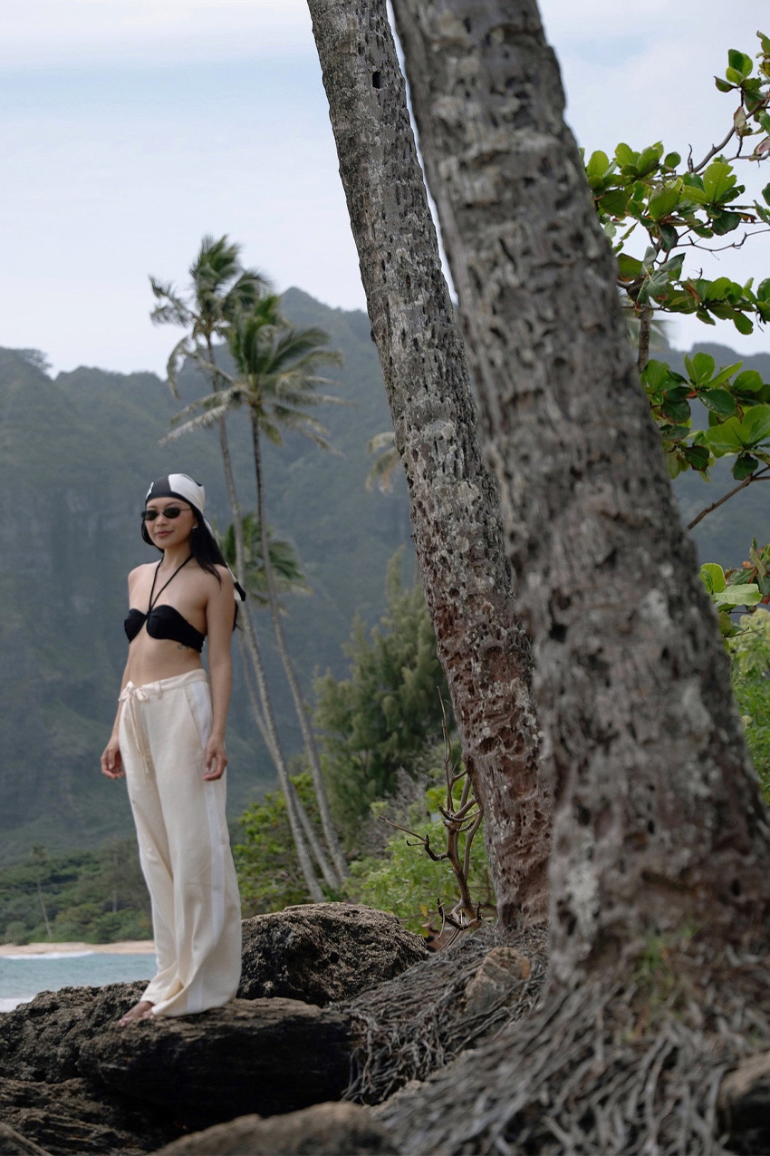 How to wear a bandana is shown in a photo of a woman standing outside by the beach wearing a printed bandana, black sunglasses, a black bikini top, and white satin trousers