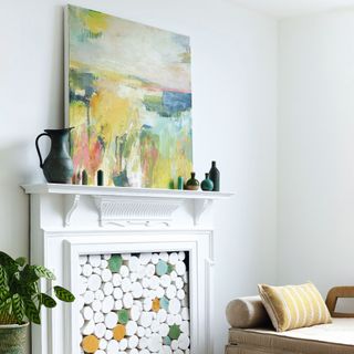 White fake fireplace with white painted logs stacked inside, large colourful canvas on mantelpiece