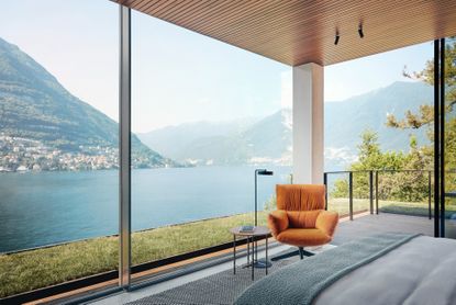 Interior of Il Sereno Lake Como Hotel with floor to ceiling windows on two sides overlooking the lake and mountains in the background. An orange armchair is visible in the room