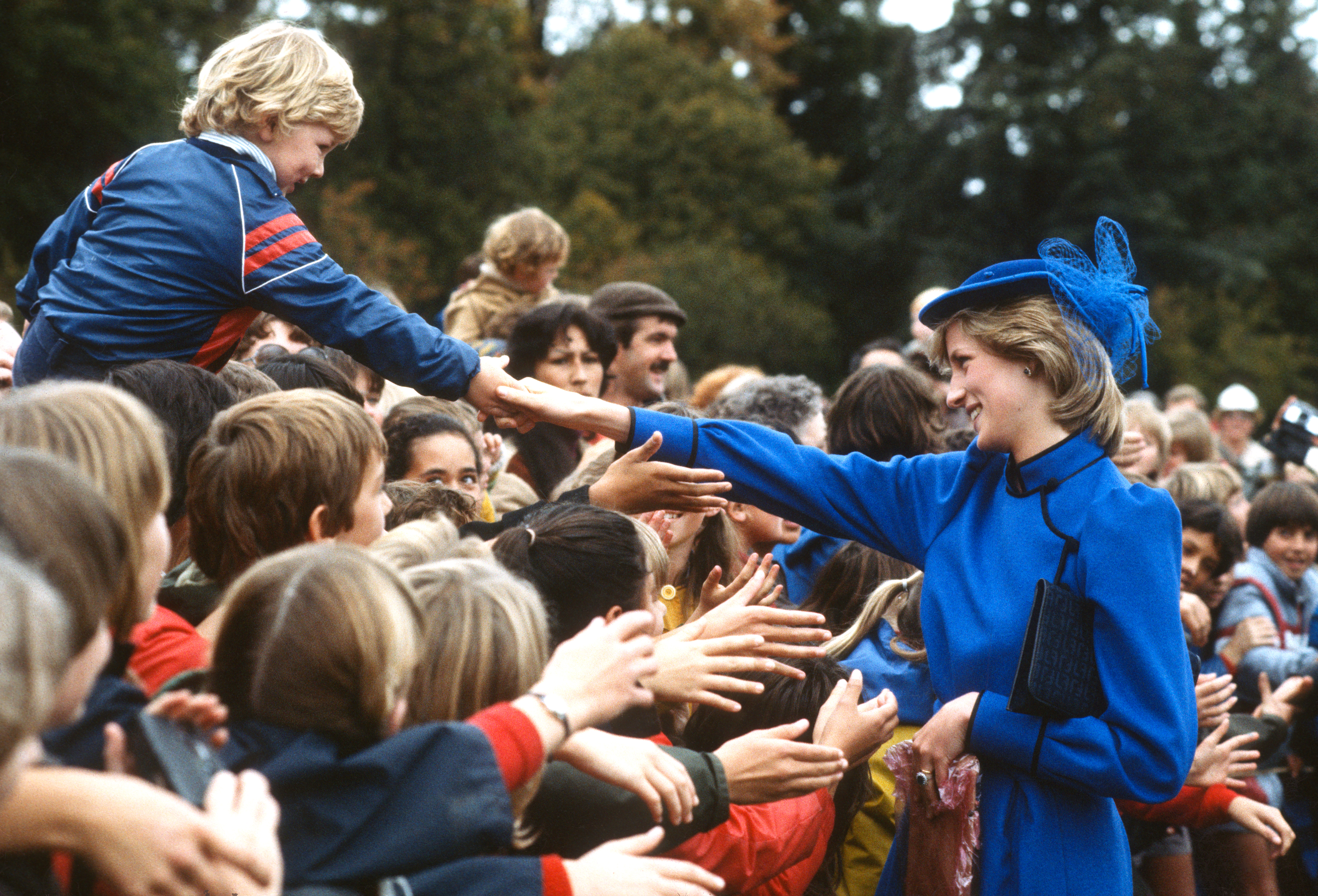 Princess Diana wearing a blue outfit and hat holding hands with a little boy in a crowd