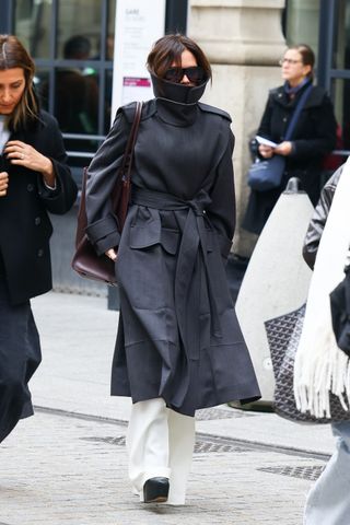 Victoria Beckham walking in Paris wearing a high neck trench coat and white flare pants with a burgundy tote bag