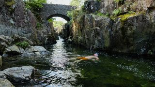 Man wild swimming in the Lake District, North East of England. He is in a river, enjoying time outdoors, floating on his back.