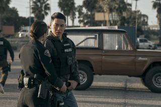 A police officer (Rich Ting as Tyson Koyoma), speaking to a female officer as they stand in front of a brown truck, in 'On Call.'