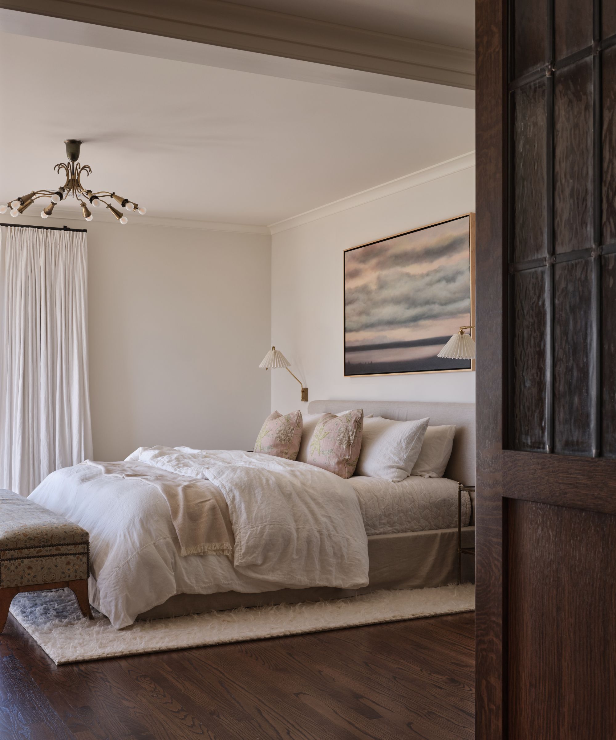 A serene bedroom with a linen-upholstered bed, white bedding, and a large landscape painting mounted between two pleated brass sconces