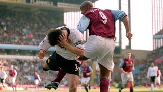 two soccer players, one in a red jersey and white shorts and one in a white jersey and black shorts, pass each other on the pitch, having just collided. The player in white holds his head in one hand as if in pain