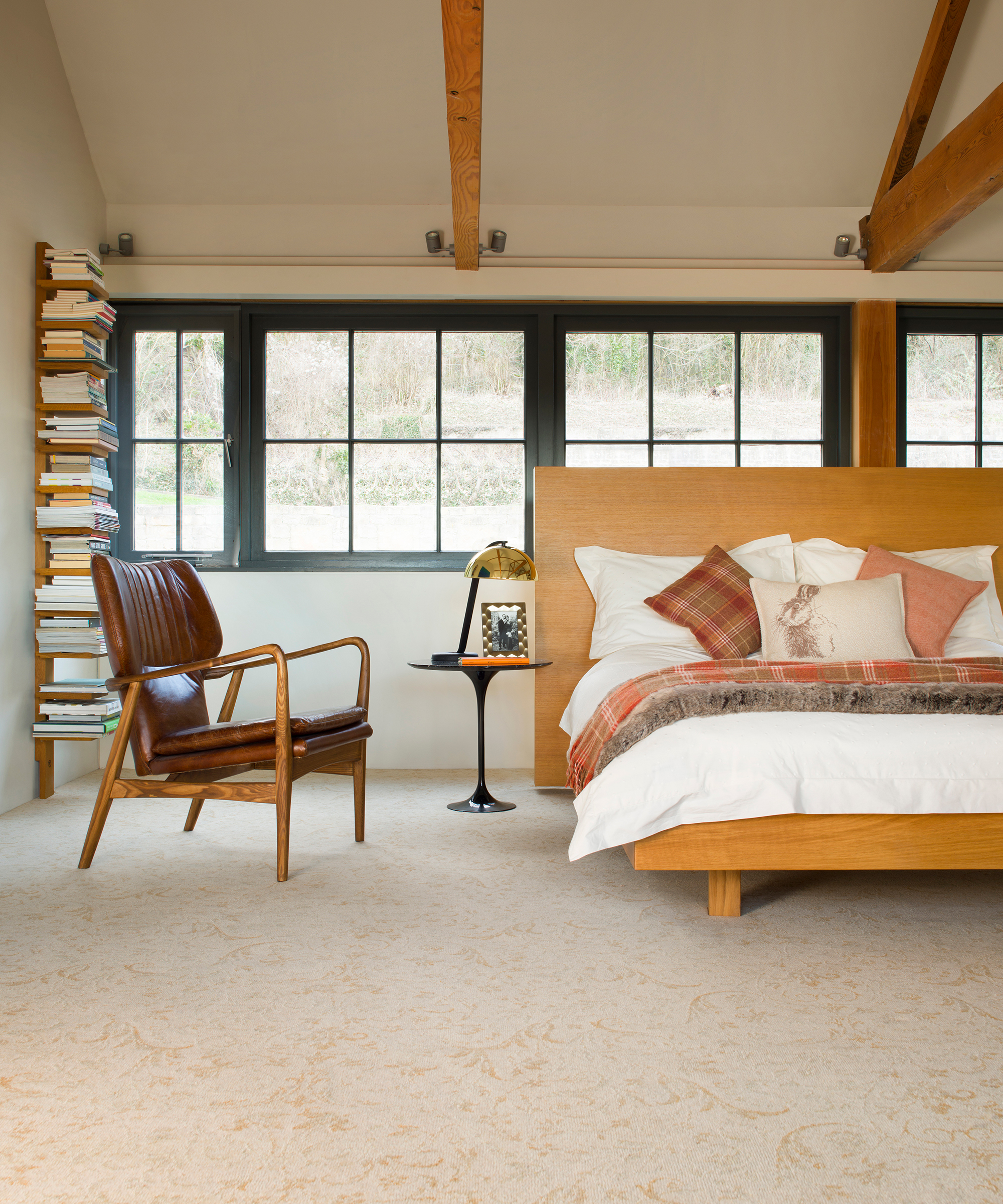 Pale gold carpet with subtle floral pattern, leather retro chair, stacked books on vertical shelf, and exaggerated wood headboard.