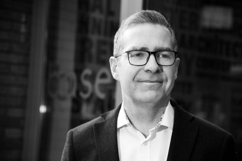 A black and white image of a man in a suit with glasses but no tie