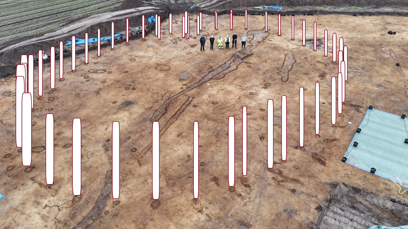 Drone-level image of a field with a ring of post holes; there are recreations of vertical timbers shown in each of the holes. Six people stand in the top center for scale.