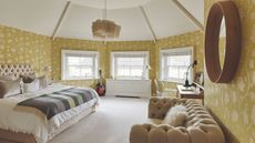 Yellow bedroom with vaulted ceiling, wooden beams painted yellow, floral wallpaper, cozy bed with throws, chesterfield sofa and desk with lamp
