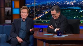 Conan sitting on the blue chairs next to Stephen Colbert's desk as both laugh on The Late Show With Stephen Colbert.