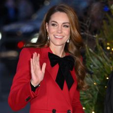 Kate Middleton wearing a red coat and a black bow waving and smiling in front of a Christmas tree