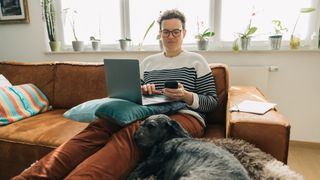 Man sitting on orange sofa with dog with a laptop and phone in hand