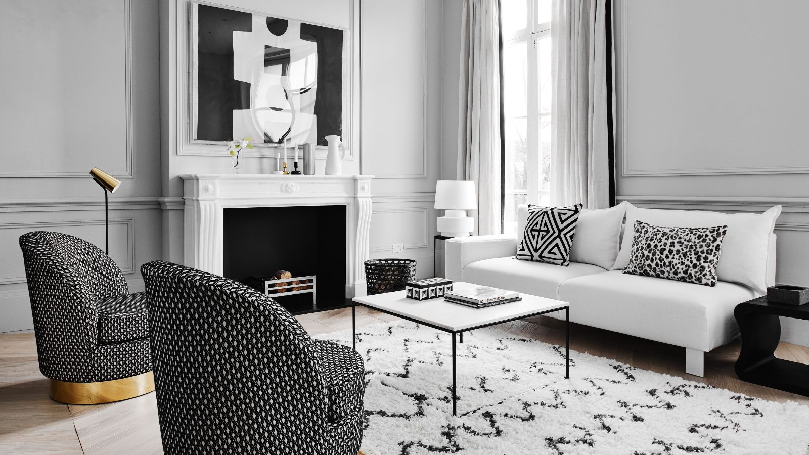 A black and white living room with white walls, a white mantelpiece, and white curtains, and a black and white artwork above the fireplace, a black and white rug, a white couch with black and white cushions, a black and white coffee table and two black and white chairs with gold bases.