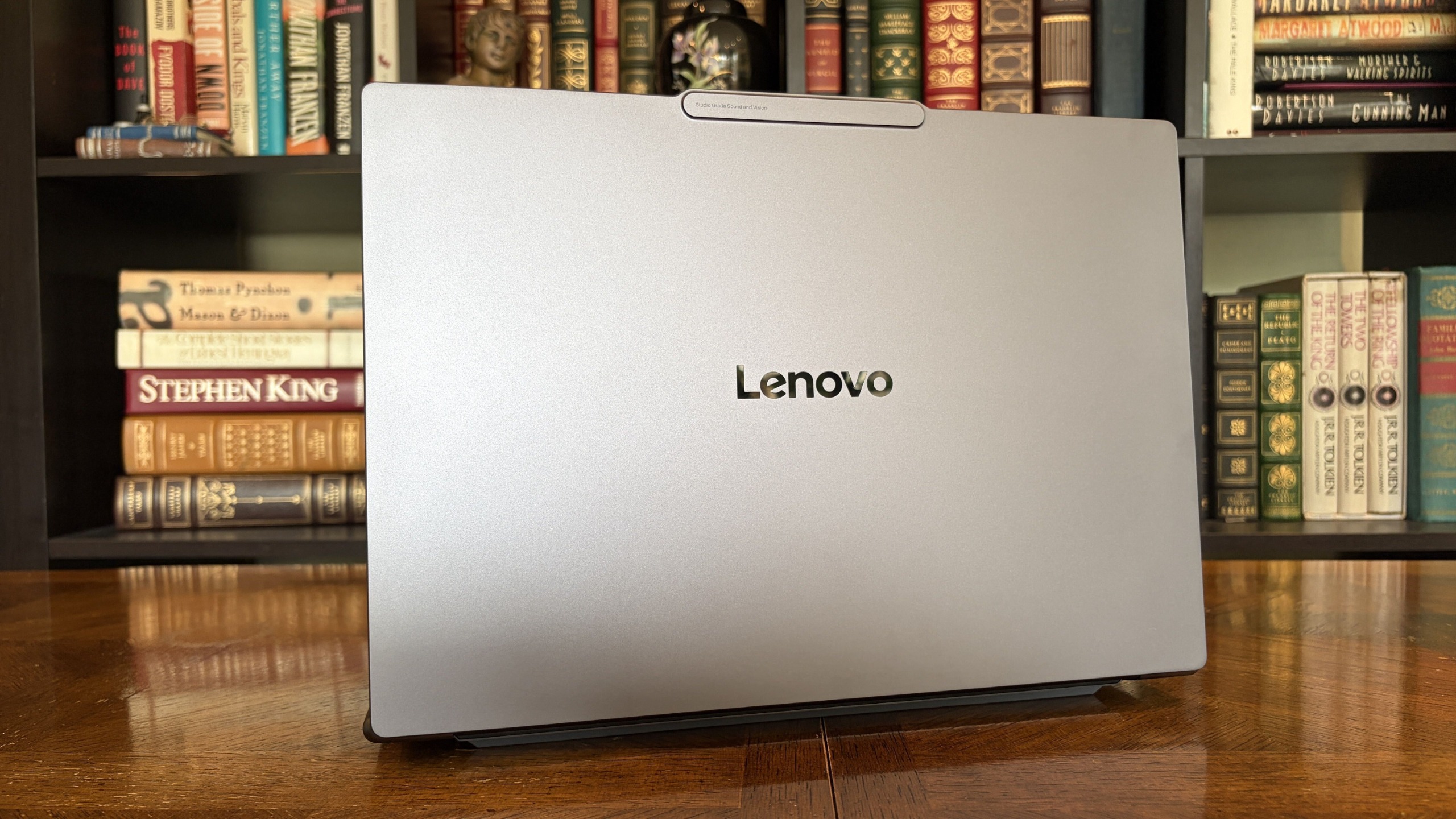A Lenovo Pro 9n laptop viewed from behind sits on a polished wooden desk before shelves of vintage books, creating a warm, scholarly atmosphere.