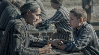 Lale (Jonah Hauer-King) tattoos Gita’s (Anna Próchniak) arm as they sit across each other at a table wearing striped gray prisoners’ uniforms in “The Tattooist of Auschwitz”