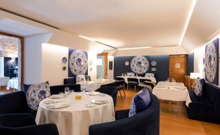 Inside look of the Ânfora restaurant. White and deep blue walls, with ornamental painting in the same color scheme. The tables are covered with white linen, and the sitting area is in deep blue velvet.