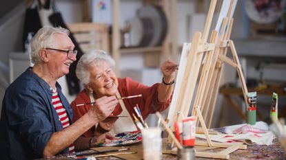 An older couple smile as the work on a painting together.