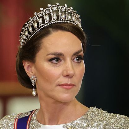 A shoulders-up photo of Kate Middleton wearing the Lover's Knot tiara and a white embellished gown with pearl earrings looking to the side with a serious expression