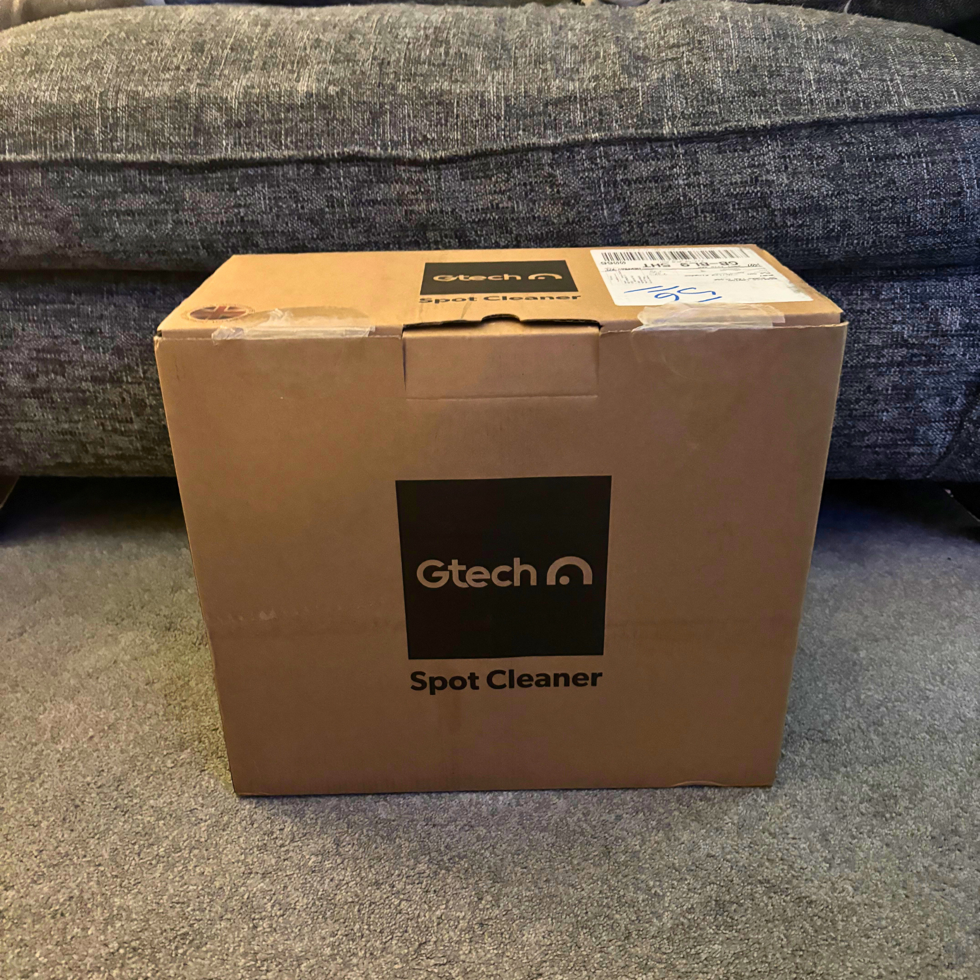 Gtech spot cleaner in brown cardboard box placed on grey carpet in front of sofa