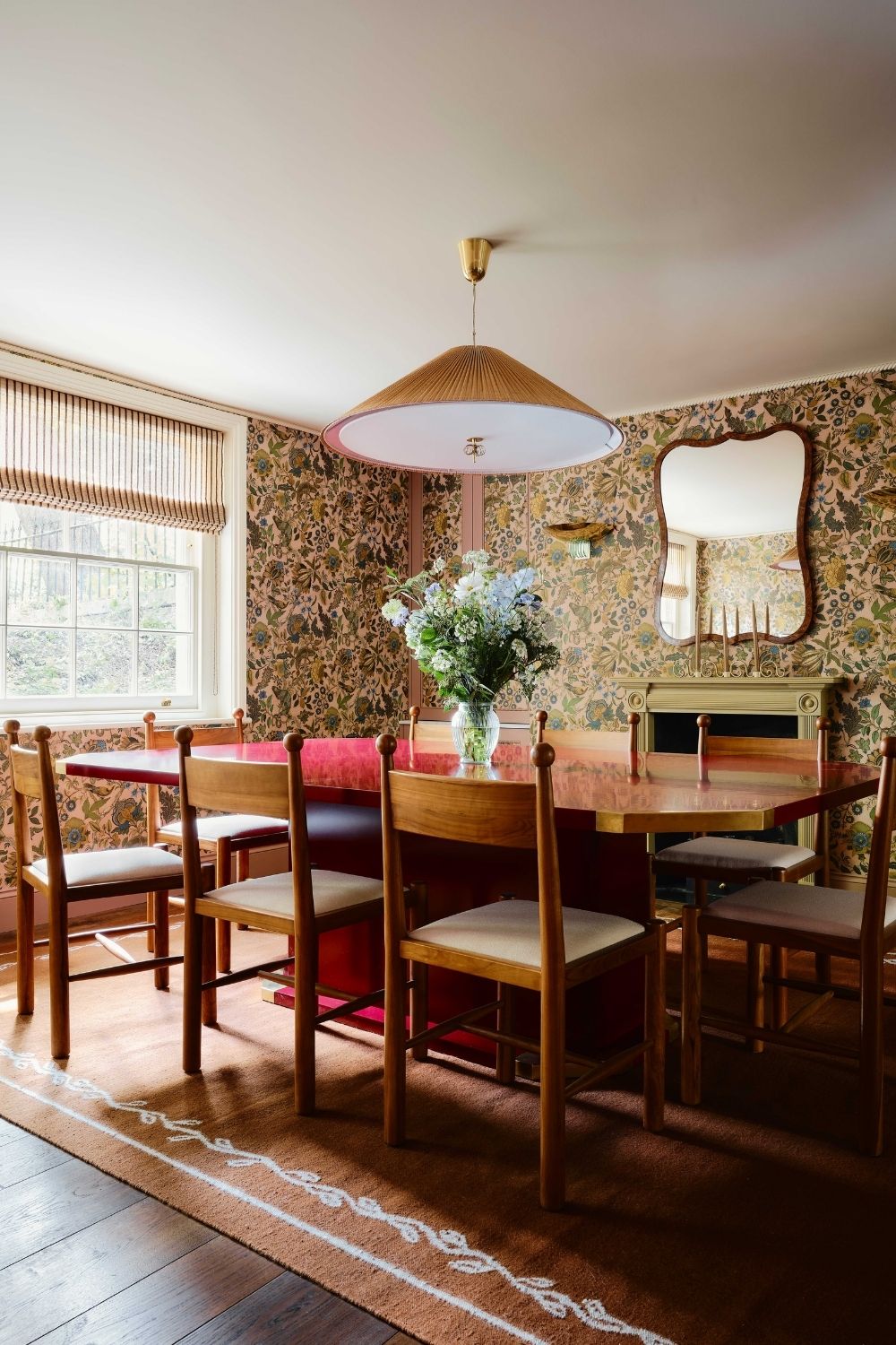 floral wallpapered dining room with a large red gloss lacquered table, wooden antique chairs with ball detailing, a large rug with border detail and a fireplace and mirror