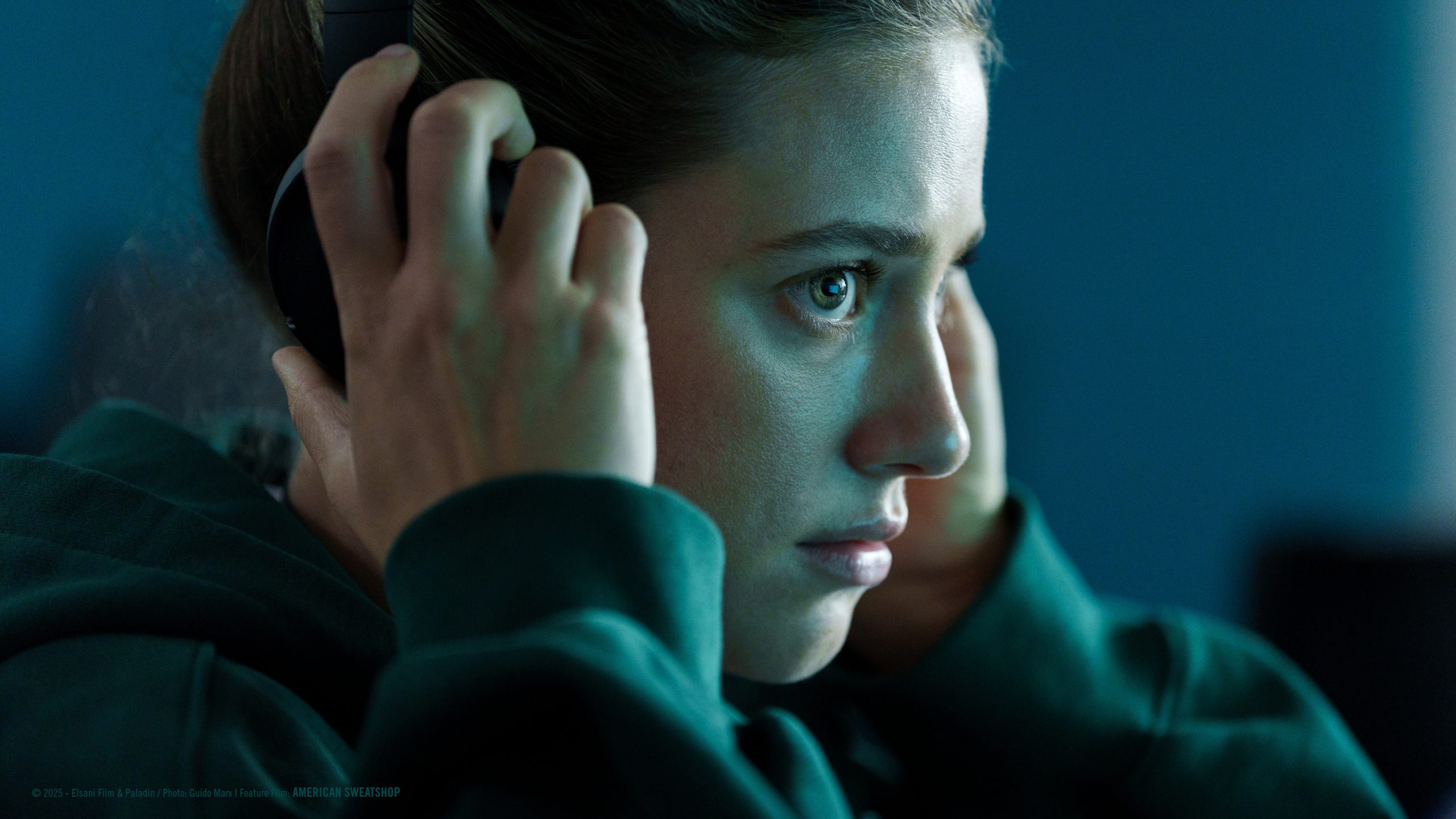lili reinhart puts on headphones while sitting at a computer in the movie american sweatshop