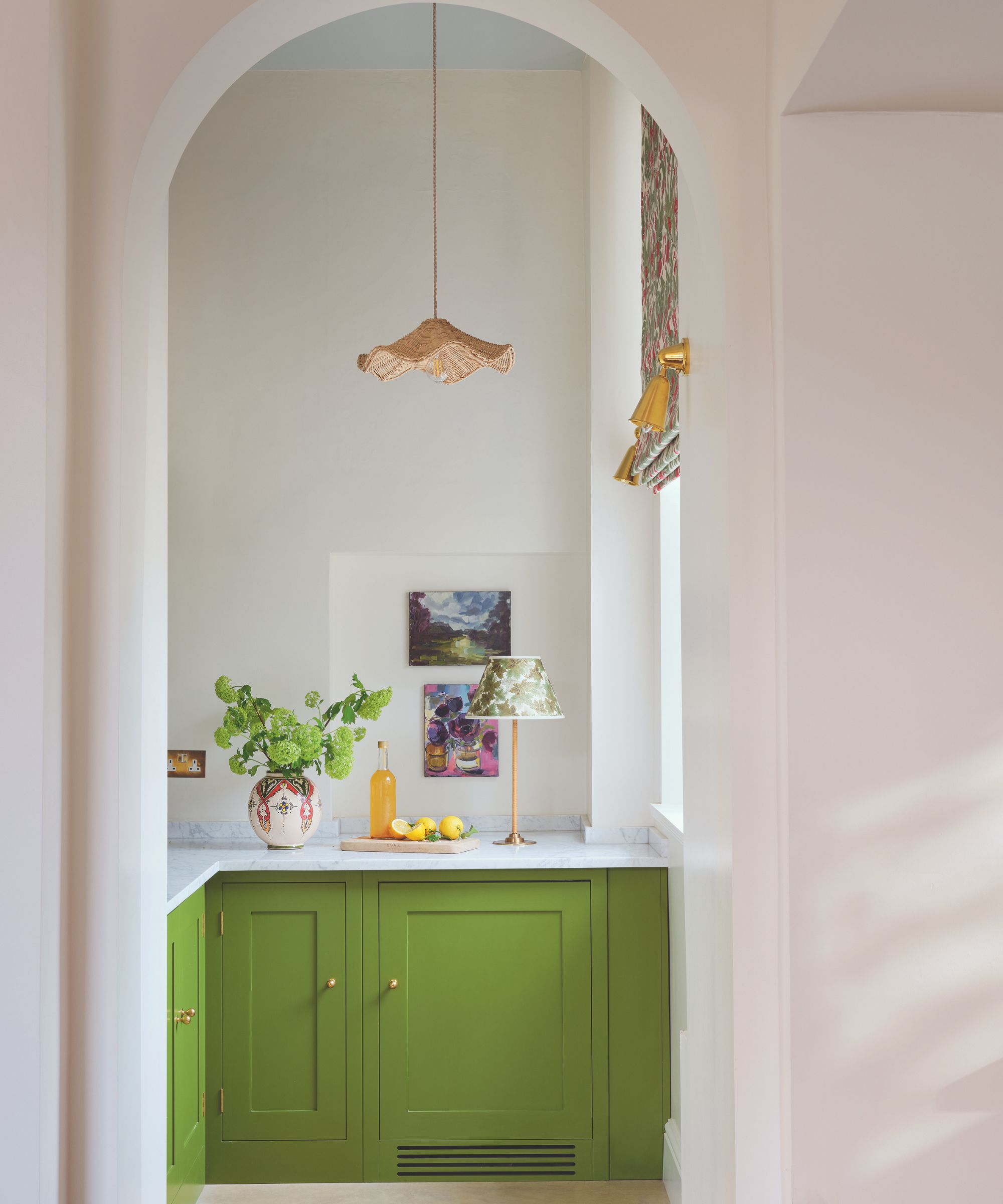 Arched doorway leading into a small kitchen with bright green cabinetry, marble countertops, window with a roman blind, a rattan pendant light, a cordless lamp on the countertop and a vase of flowers