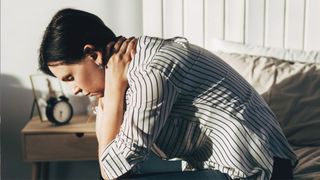 The image shows a dark haired woman wearing a stripy shirt sitting on the edge of a bed and holding her head in her hands