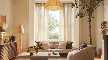 Image of an off-white living room with a large window decorated with linen curtains. There is a low sofa and an accent chair with a rectangular coffee table in between.