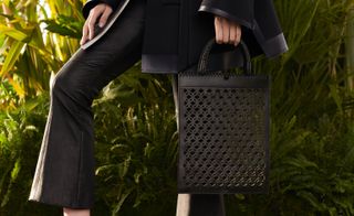 View of a model dressed in a black jacket and black flared trousers holding a black handbag with a cut-out, transparent and geometric design. The model is standing in front of greenery