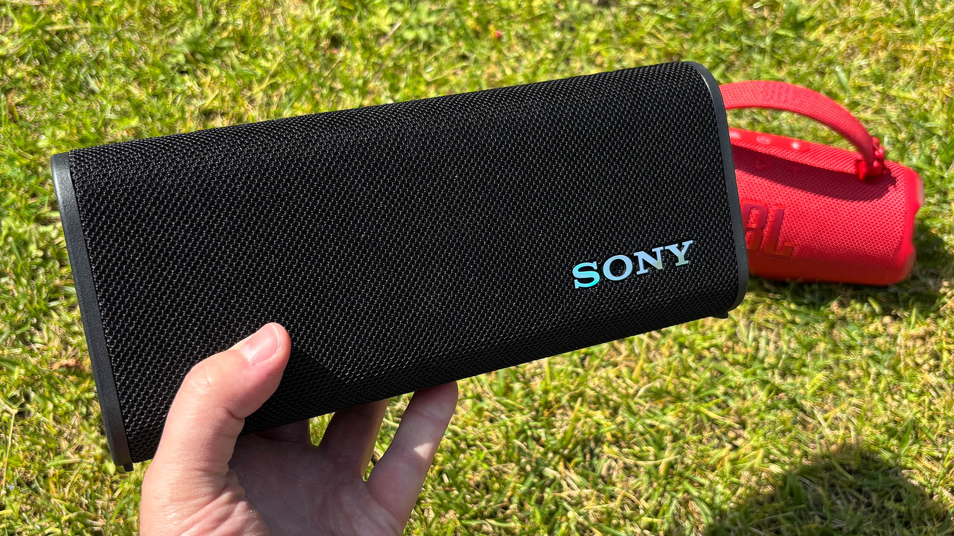 Sony ULT Field 3 wireless speaker held in hand over grassy lawn with red JBL Charge speaker behind it