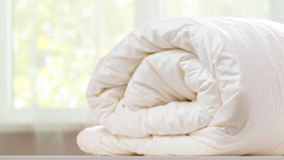 A white wool duvet rolled up on a bed with a window in the background showing a sunny day