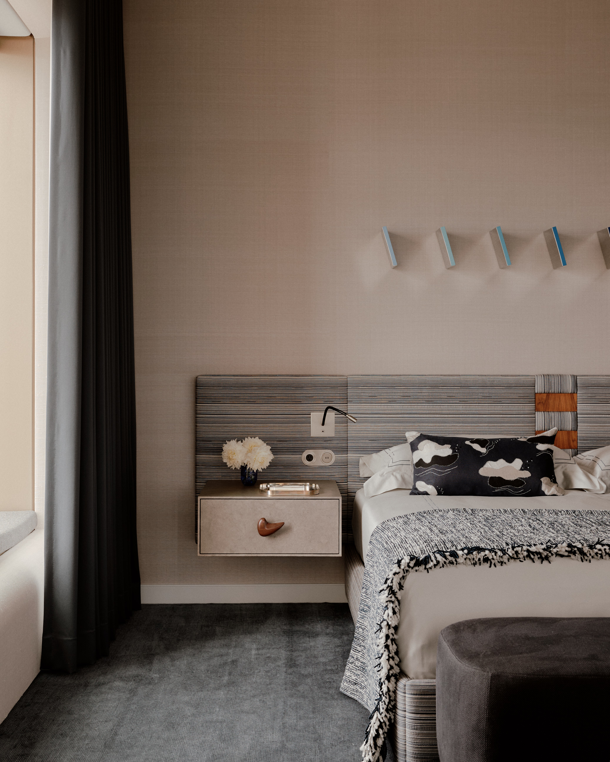modern bedroom with dark carpet, dark curtains, beige wall, built-in headboard with striped upholstered, neutral bedding, ottoman at the end of the bed, and sculptures hanging above it