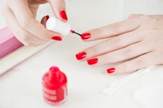 A close up of a woman's hands painting her nails win a birght pink colour with a nail brush. The bottle of nail polish is next to her hands alongside some cotton buds on a white towel.