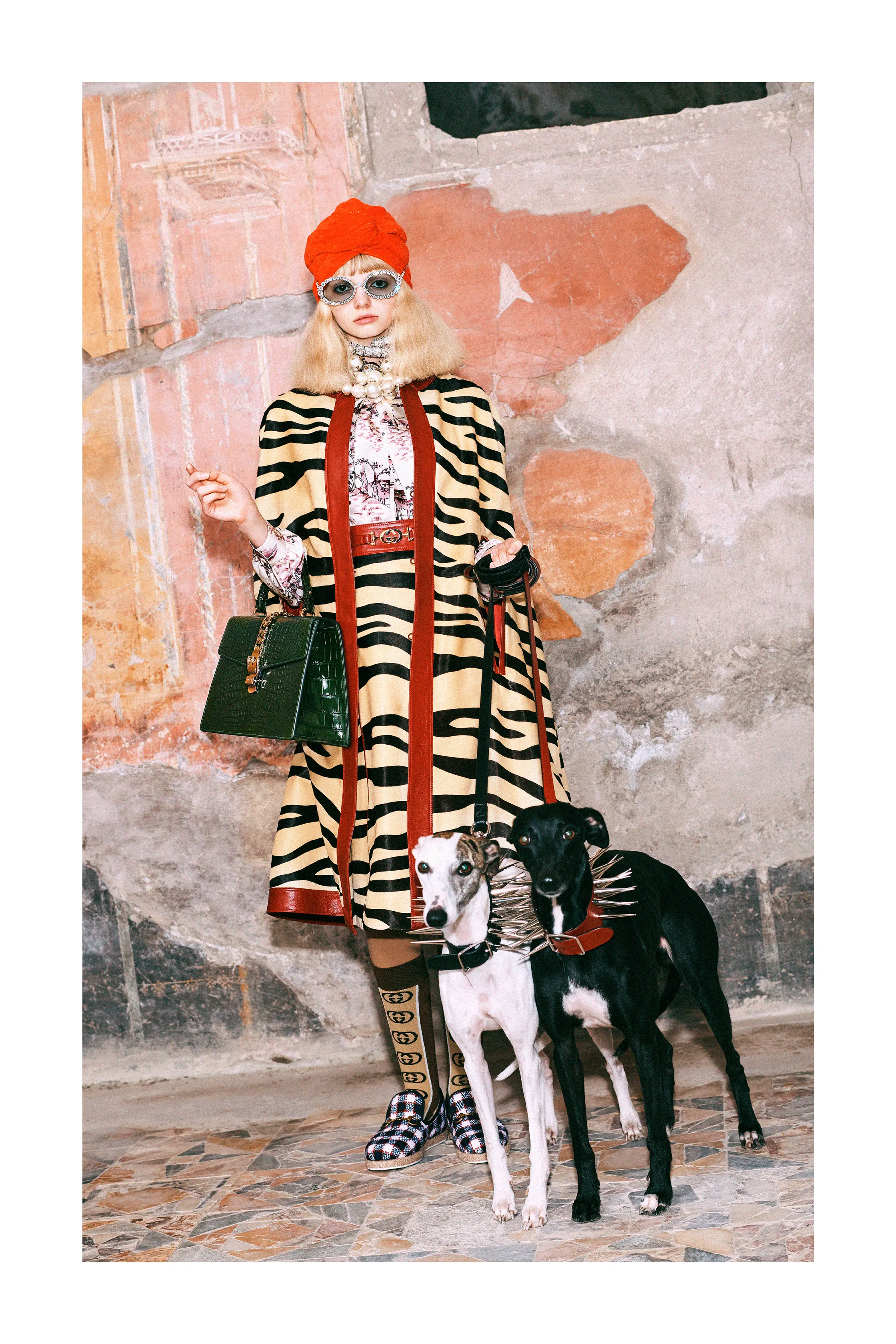 A model at Gucci Pre-Fall 2018 wearing a tan and black tiger print coat and skirt, white and pink blouse, red turban, black and white socks, with two dogs