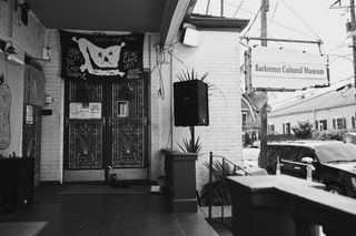 A black and white photo of a museum entrance features a boxy sign that reads "Backstreet Cultural Museum", speakers, and an ornate iron door decorated with a textile depiction of a skull.