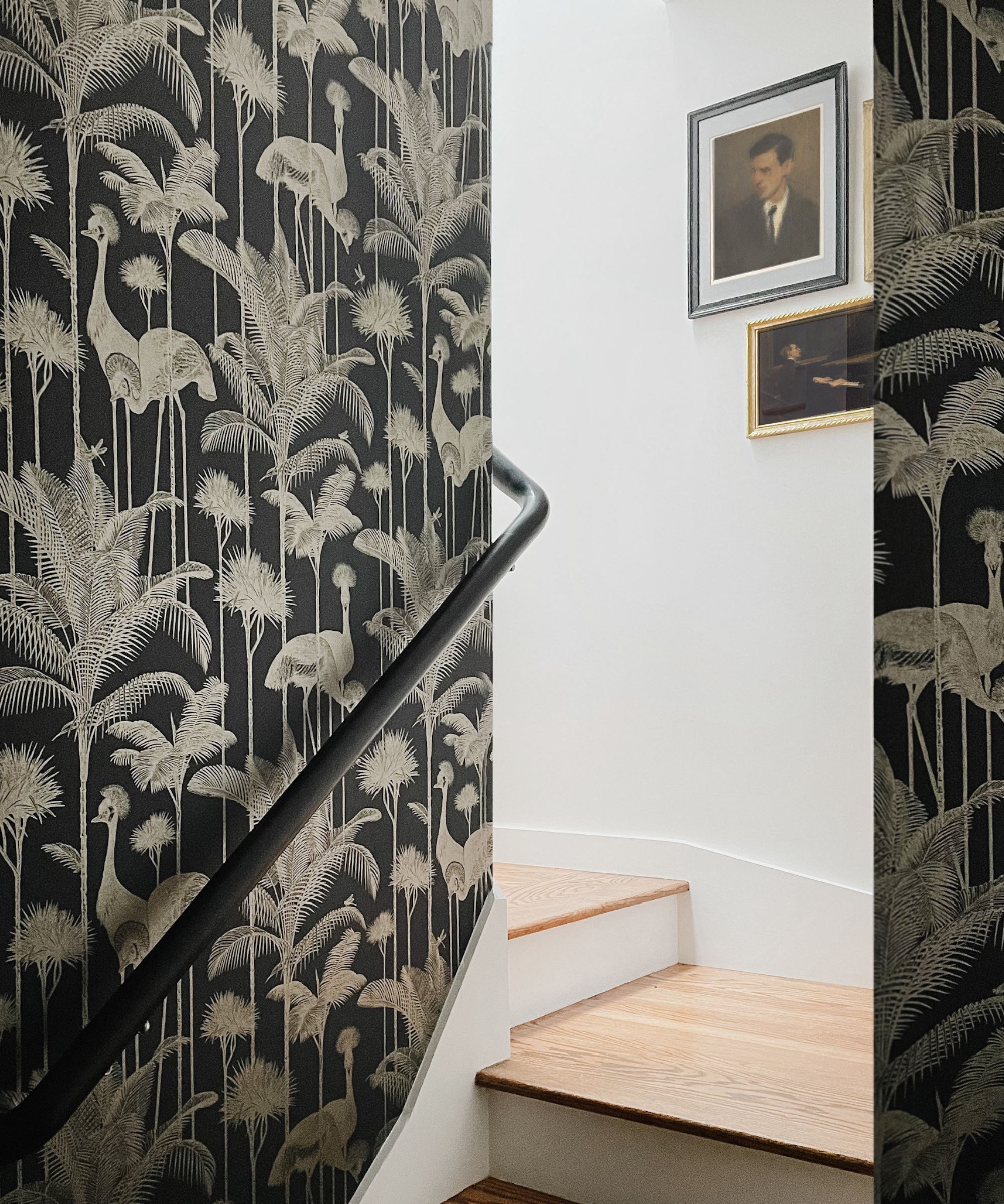 A stairwell with black wallpaper featuring tall palms and cranes in a gold outline. The stairs are pale wood with white walls above, decorated with framed portraits.