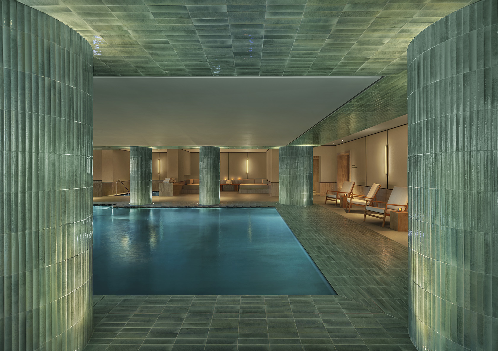 A turquoise stone-clad subterranean spa with chunky pillars covered in tiles made of the material and reclining loungers in pale wood and beige fabric, all illuminated by suffused lighting.