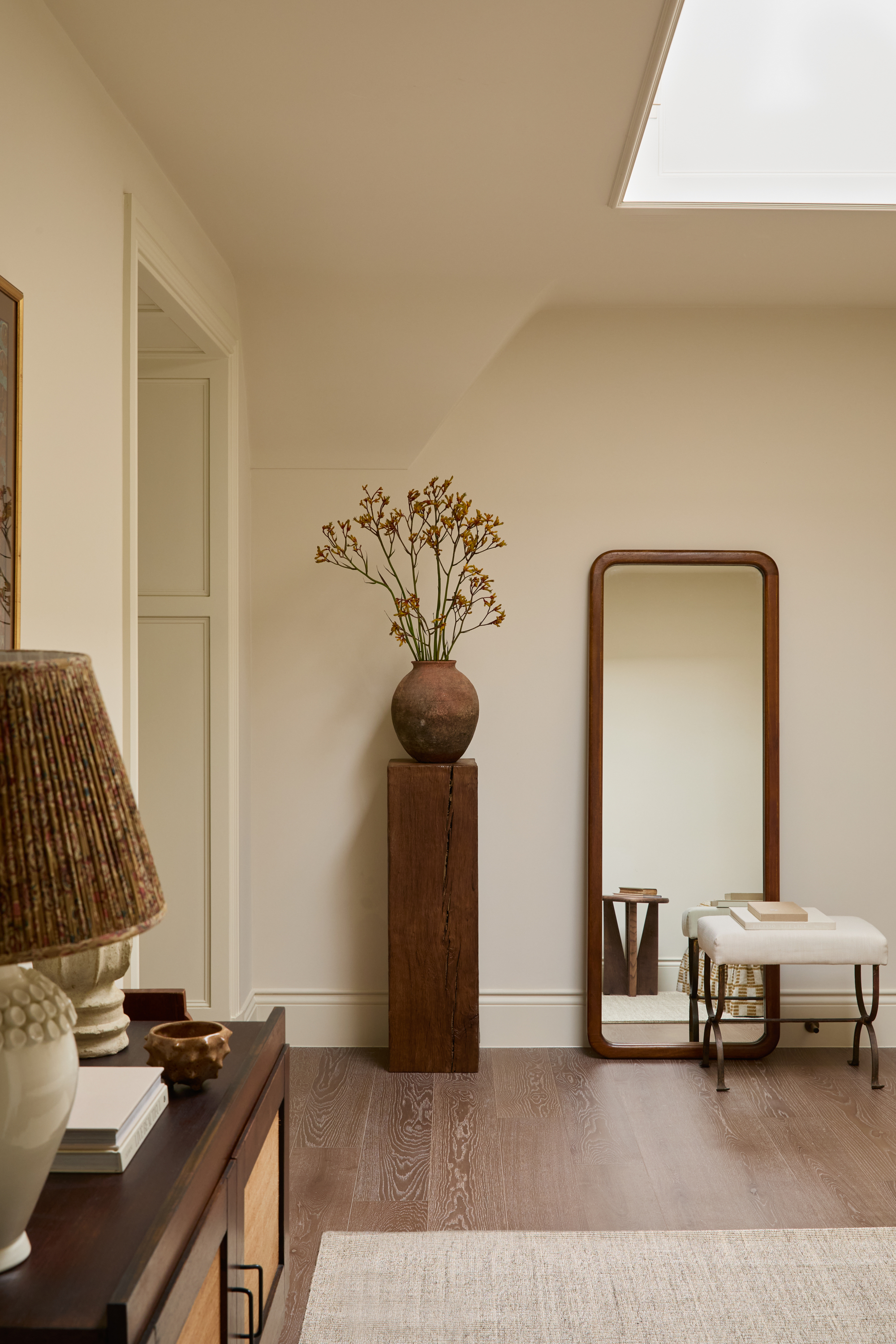 neutral hallway with timber plinth holding terracotta vase with flowers, leaning full-length mirror with ottoman in front, console with lamp, books, and vases, and woven rug on timber floors