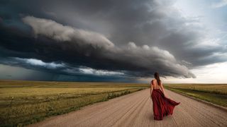 Woman in a red dress walking down a rural road under an ominous stormy sky