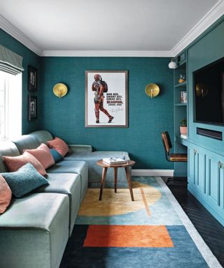A teal blue living room with a gray velvet couch with pink and blue throw pillows, a colorful blue orange and yellow carpet, a TV on the right wall and gold wall sconces and a Deadpool wall art print on the centre wall