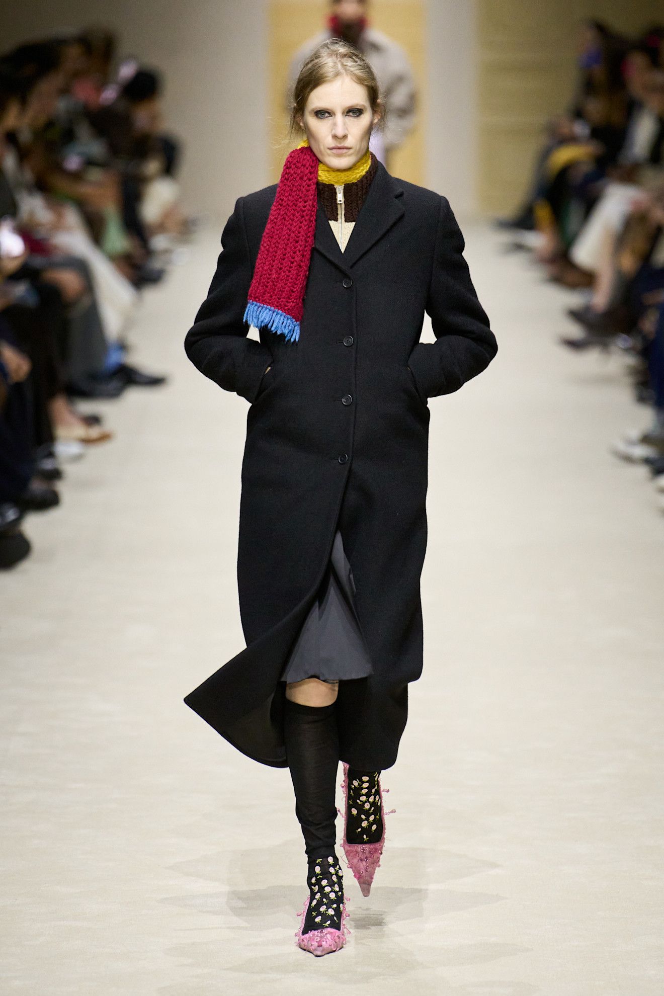A runway model wears a long black coat, black embellished socks, and pink embellished heels.