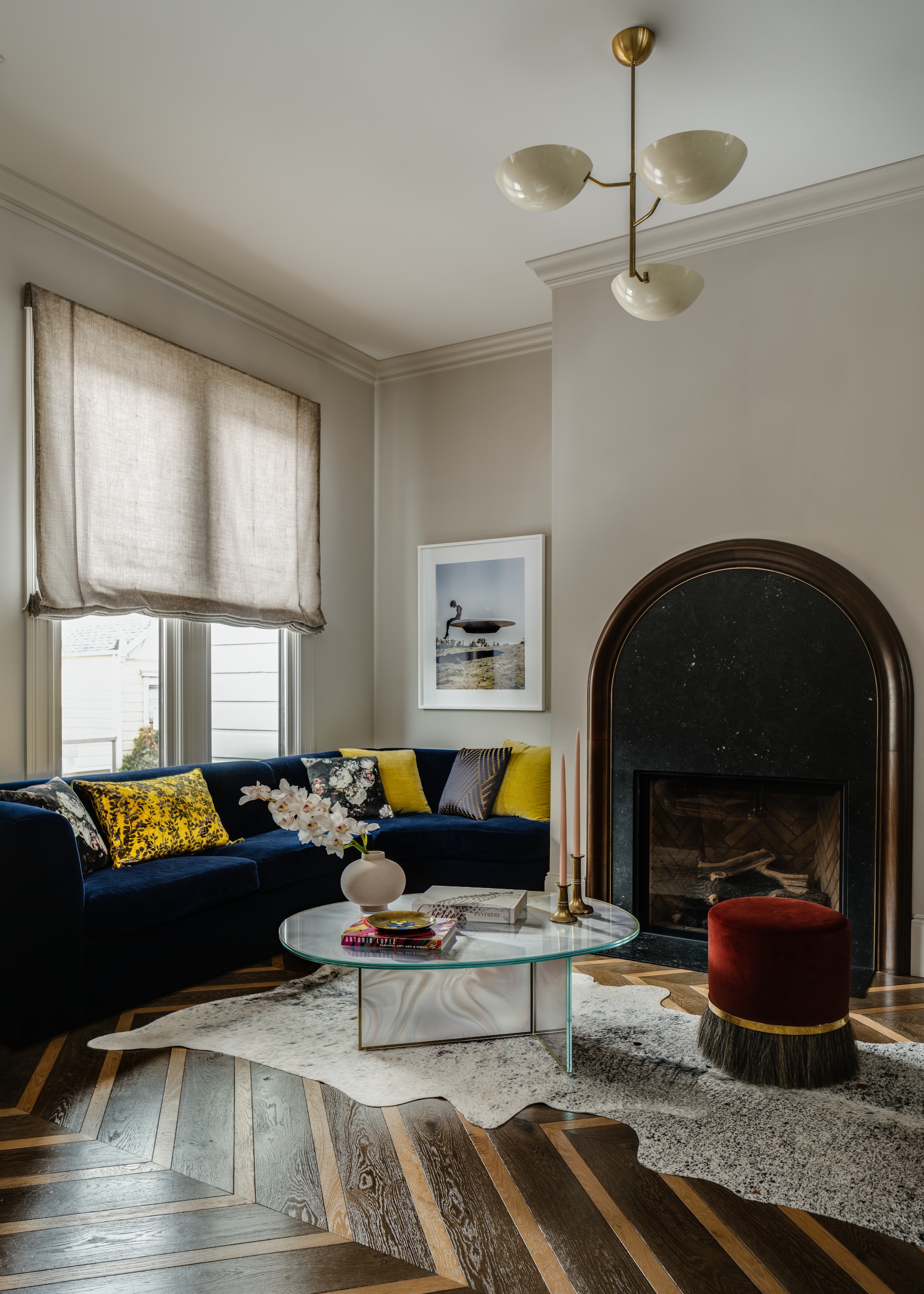 A warm taupe living room with high ceilings, large arched fireplace, blue velvet sofa and red pouffe.