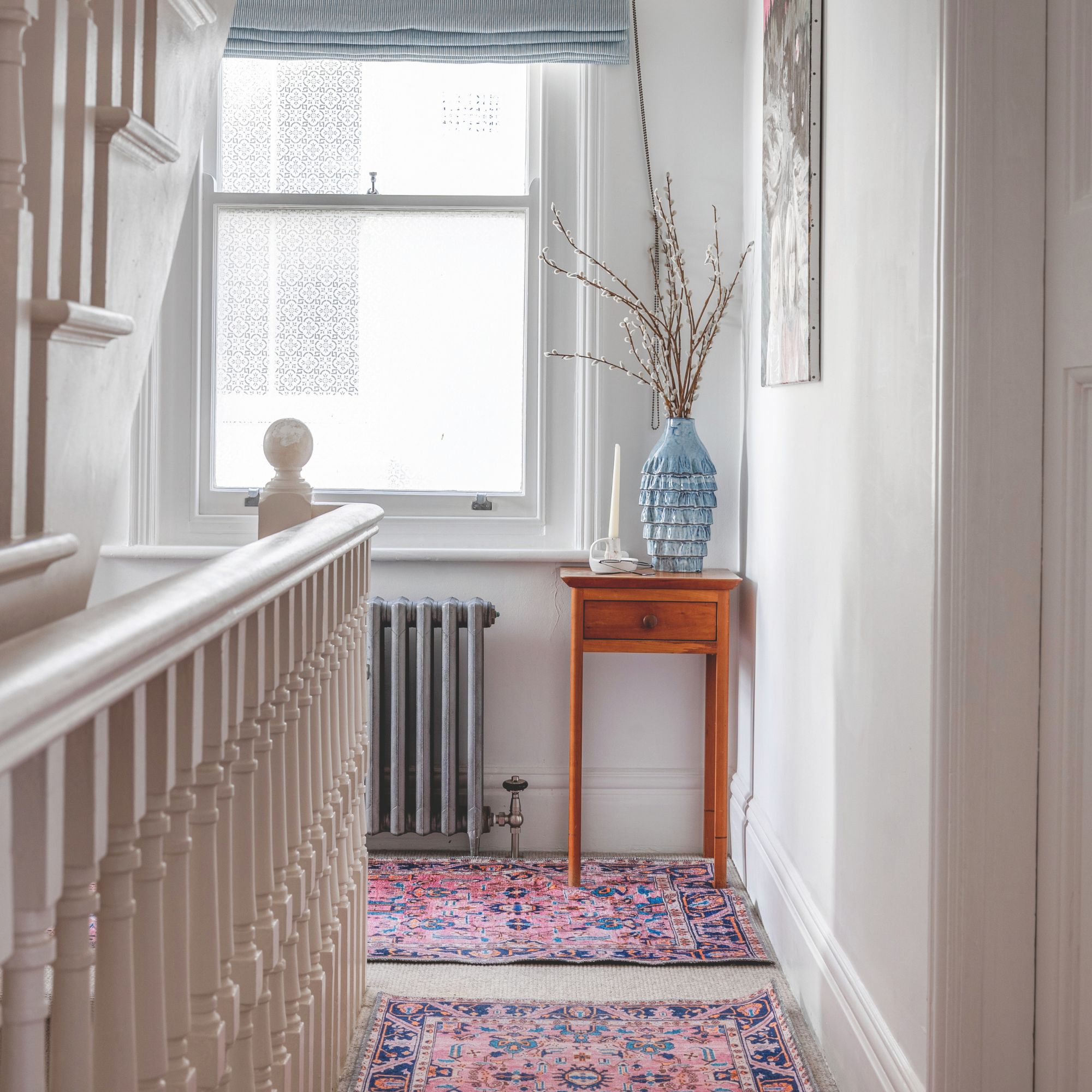 White painted landing with pink rugs on the floor and a side table with a vase on it