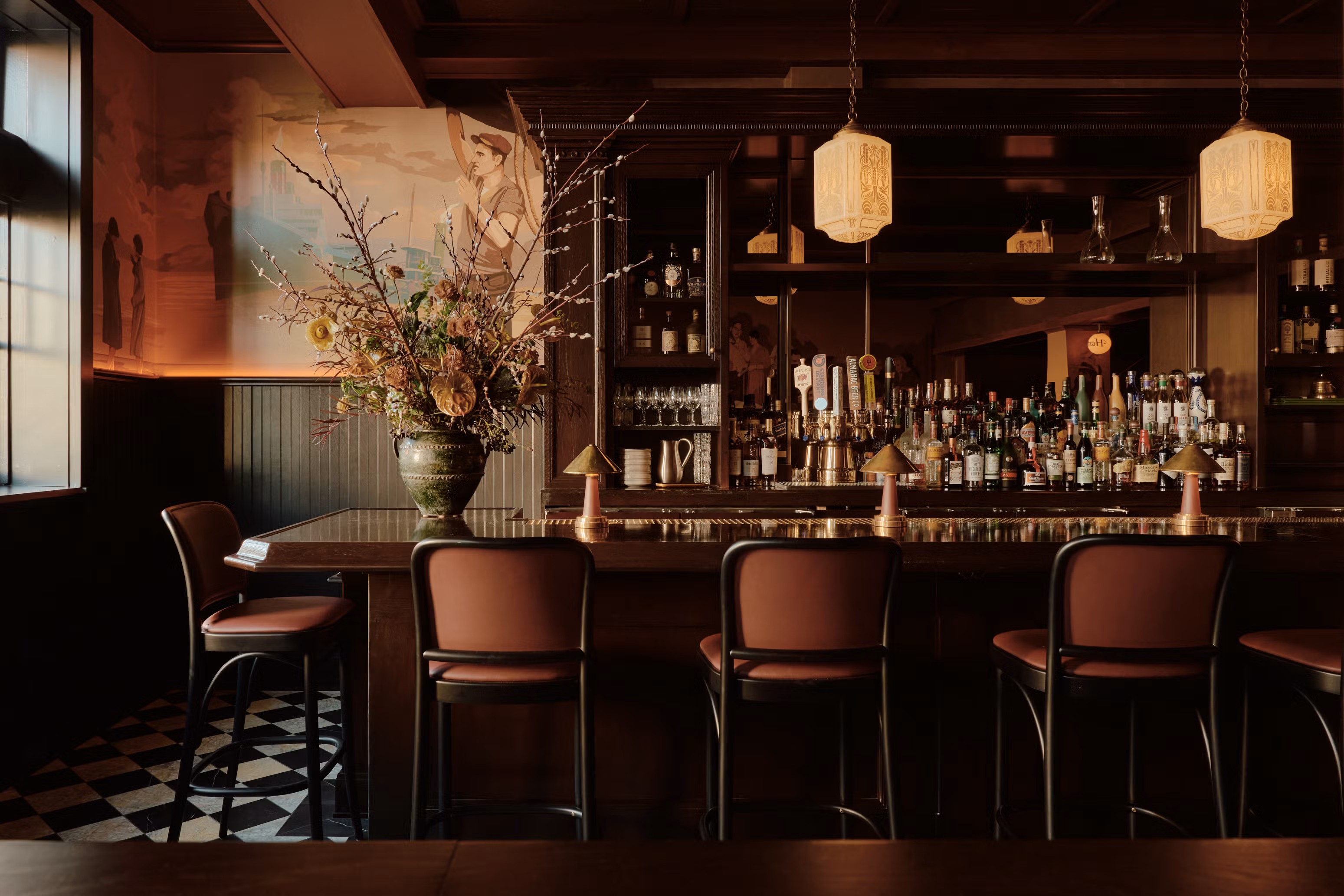 A retro-inspired, checker-floored restaurant with earthy interiors in tones of argilla, brown, orange, and light blue boasts a lacquered bar counter with mushroom-shpaed copper tablelamps, leather and wood red and black stools, a crafty ceramic vase filled with flowers in green, and handpainted murals.