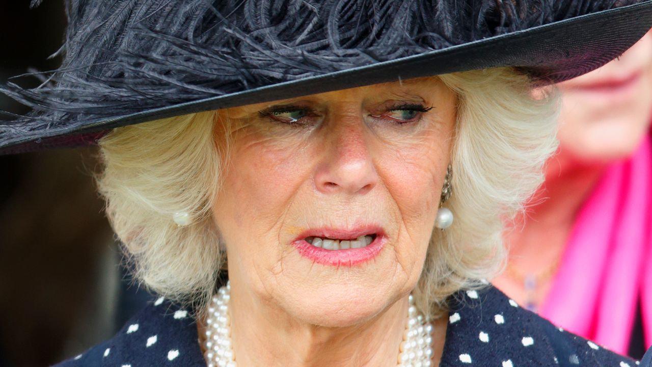 Queen Camilla wears a polka dot dress, a pearl necklace and black hat with feathers in it and she looks tearful