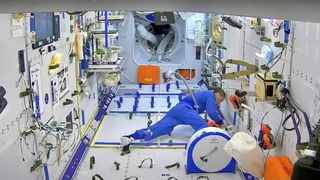 an astronaut in a blue flight suit cleans the inside of a white space station room with a small vacuum.
