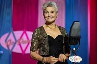 The Dancing Duchess (Angela Rippon) stands on the set of the fictional TV show Go Dancing!, with a 1950s-style stage microphone on a stand in front of her. She is wearing a black evening dress with a sequinned jacket over the top. The backdrop behind her has vertical strips of blue, magenta and pink alternating.