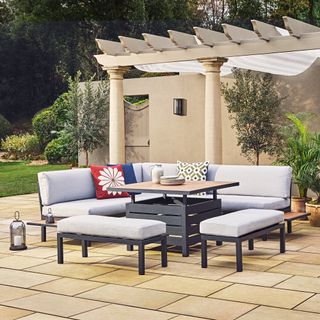 Black framed corner sofa and benches with light grey cushions and an adjustable height table for dining on a paved patio with pergola behind