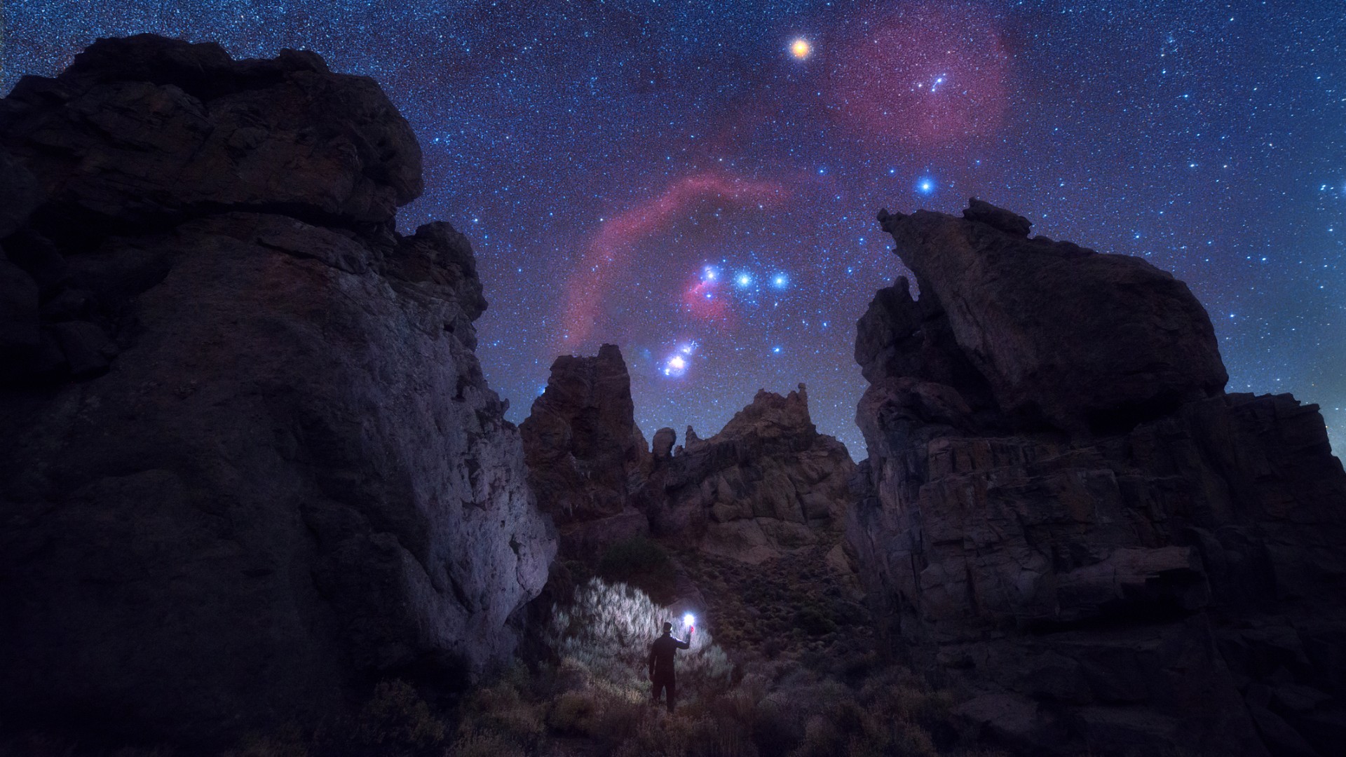 A night sky showing the constellation Orion peaking between large craggy rocks. At the bottom is a human holding up a light.