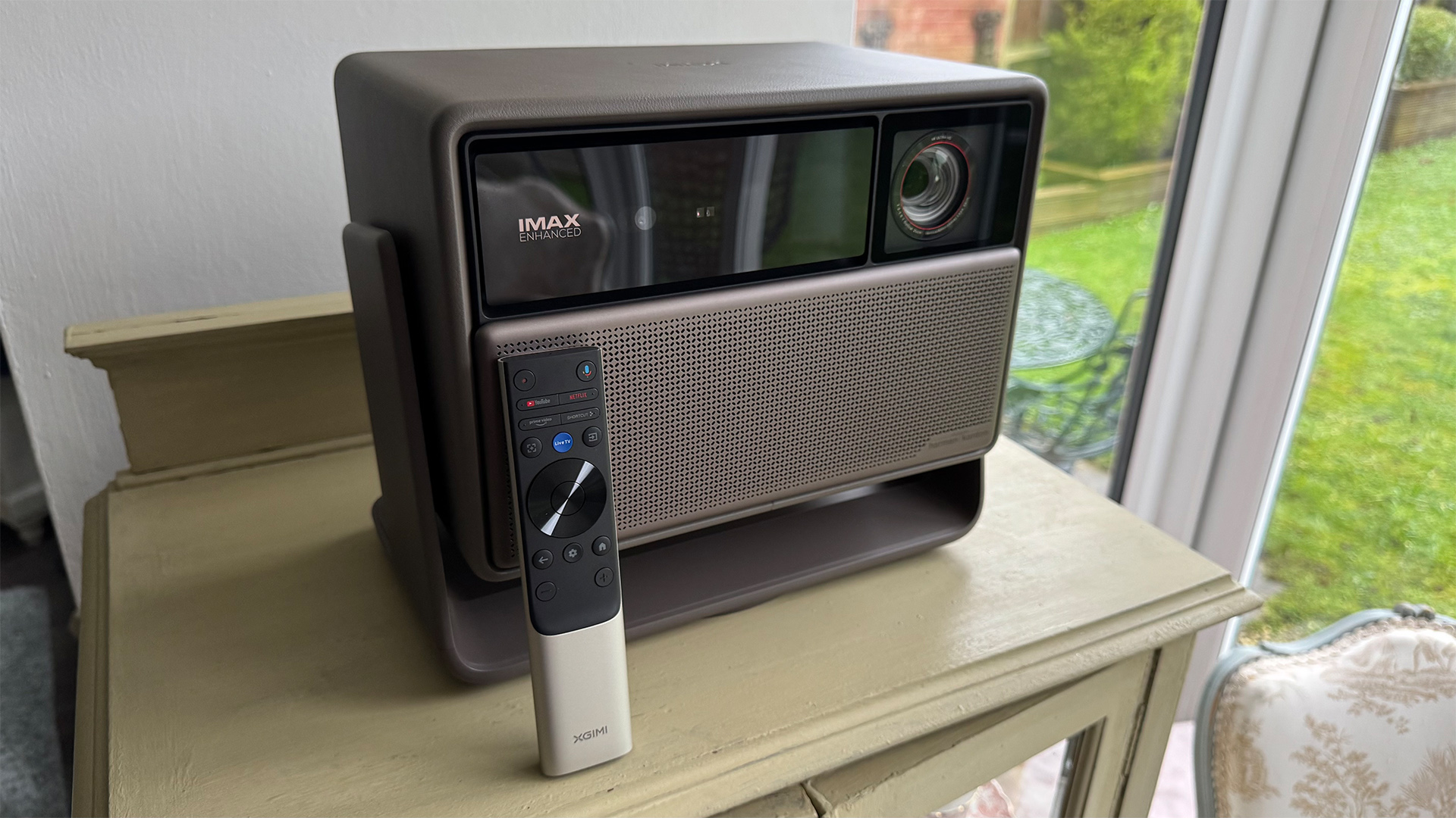 Xgimi Horizon 20 Max home cinema projector on wooden furniture next to window with remote control in front of it